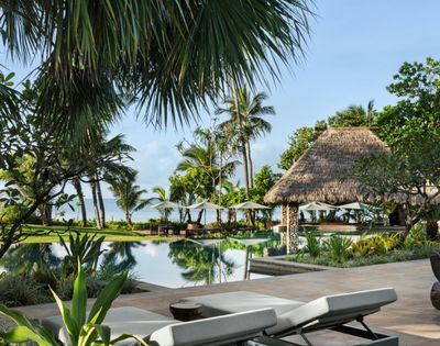 Tropical resort scene with a tranquil pool surrounded by lush palm trees and a thatched-roof gazebo. Lounge chairs face the calm ocean.