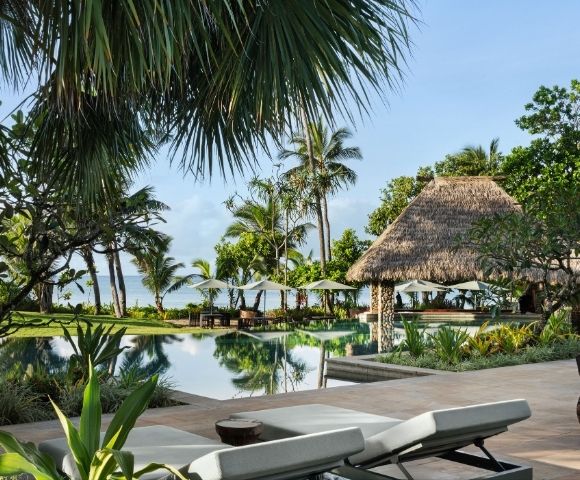 Tropical resort scene with a tranquil pool surrounded by lush palm trees and a thatched-roof gazebo. Lounge chairs face the calm ocean.