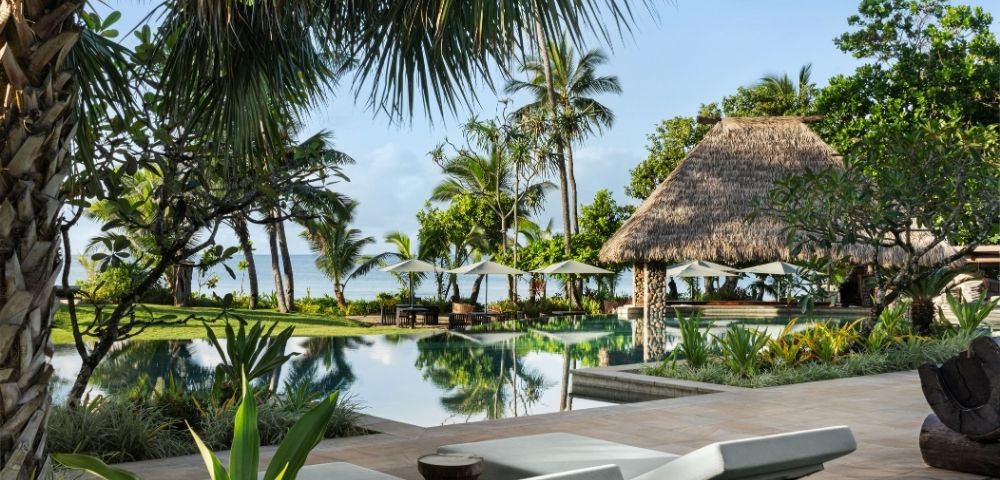 Tropical resort scene with a tranquil pool surrounded by lush palm trees and a thatched-roof gazebo. Lounge chairs face the calm ocean.