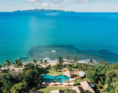 Aerial view of a tropical beach resort with clear turquoise water, palm trees, a lush garden, and a large pool surrounded by sun loungers and thatched huts.