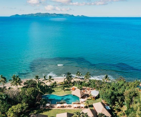 Aerial view of a tropical beach resort with clear turquoise water, palm trees, a lush garden, and a large pool surrounded by sun loungers and thatched huts.