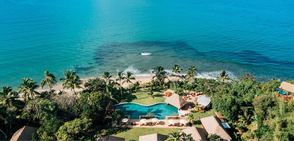 Aerial view of a tropical beach resort with clear turquoise water, palm trees, a lush garden, and a large pool surrounded by sun loungers and thatched huts.