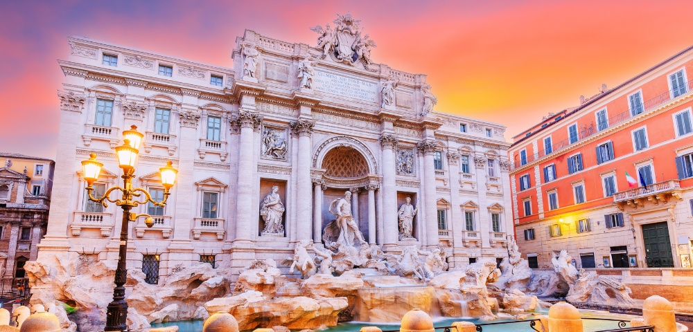 A majestic view of the Trevi Fountain in Rome at sunset, with its ornate sculptures and flowing water, illuminated by warm streetlights under a vibrant pink-orange sky.