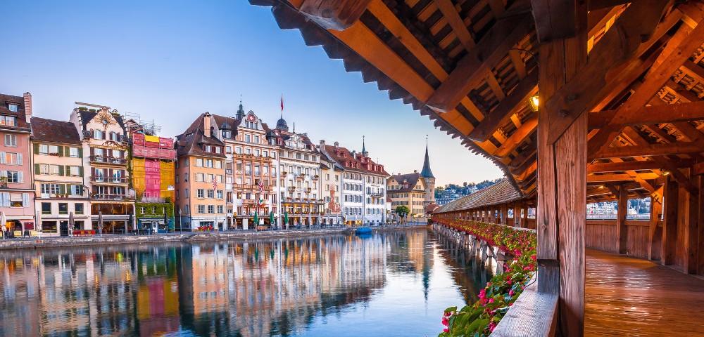 Covered wooden bridge adorned with flowers over a calm river, reflecting colorful historic buildings under a clear sky. Serene and picturesque setting.