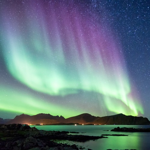 Vibrant northern lights in green and purple cascade over a serene coastal landscape with dark mountains, a starry night sky, and calm water reflecting the colors.