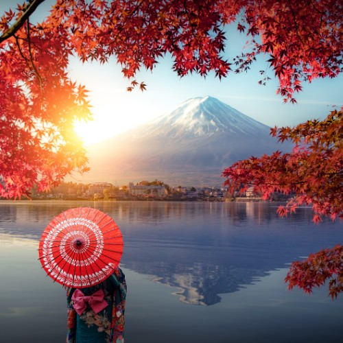 A person in traditional Japanese clothing with a red parasol stands by a lake, admiring Mount Fuji. Autumn leaves frame the serene scene.