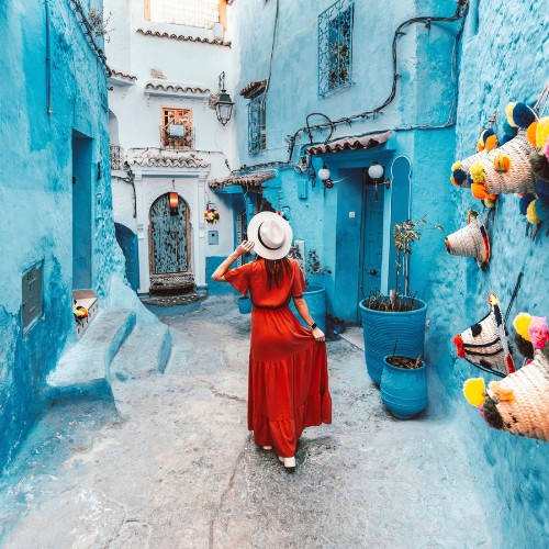 A woman in a red dress and white hat walks through a narrow, vibrant blue alley adorned with colorful woven baskets and pots, evoking a sense of wanderlust.