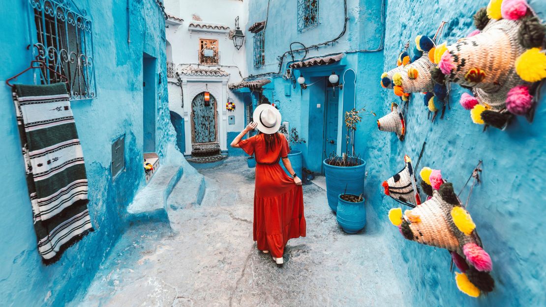 A woman in a red dress and white hat walks through a narrow, vibrant blue alley adorned with colorful woven baskets and pots, evoking a sense of wanderlust.