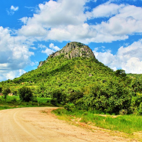 Dirt road leading to a lush, green hill under a bright blue sky with scattered white clouds. The scene exudes tranquility and natural beauty.