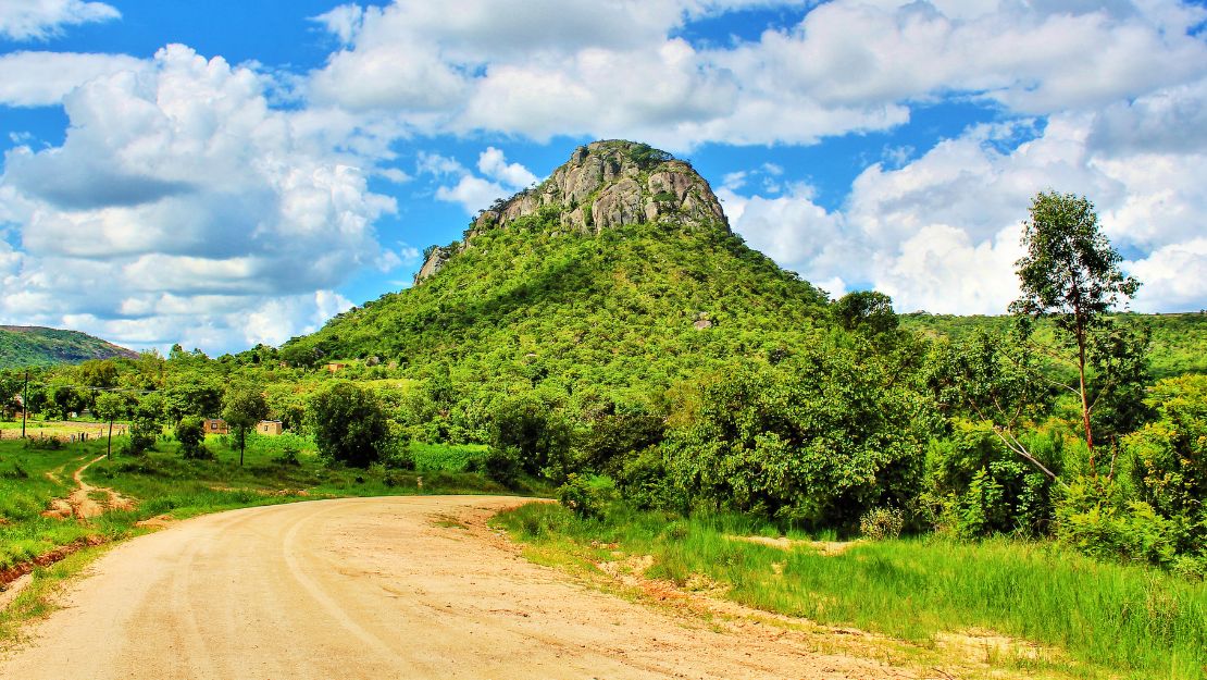 Dirt road leading to a lush, green hill under a bright blue sky with scattered white clouds. The scene exudes tranquility and natural beauty.