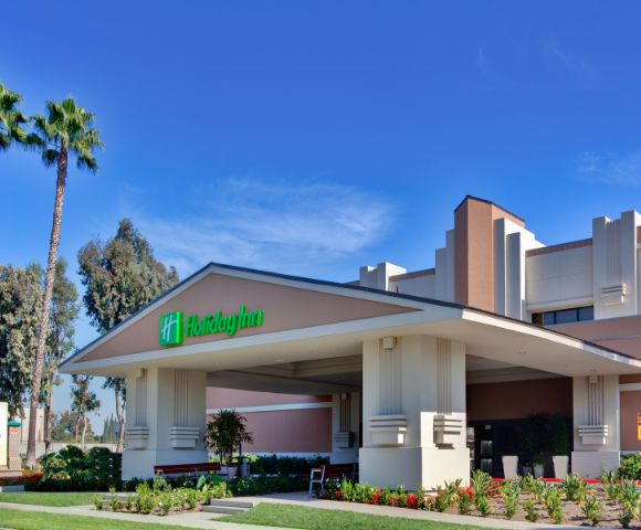 Front view of a Holiday Inn hotel entrance surrounded by lush greenery and tall palm trees under a clear blue sky, conveying a welcoming and relaxed atmosphere.
