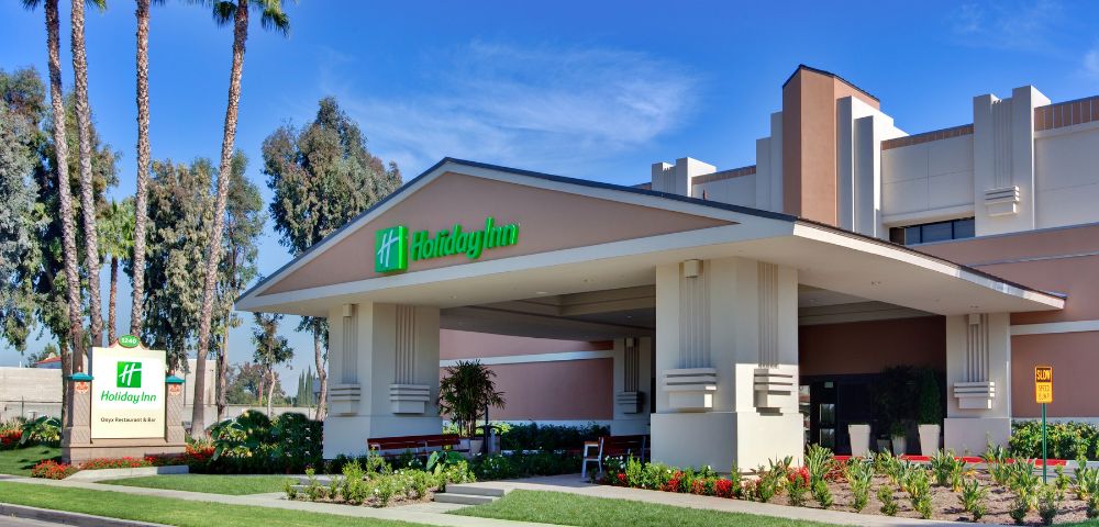 Front view of a Holiday Inn hotel entrance surrounded by lush greenery and tall palm trees under a clear blue sky, conveying a welcoming and relaxed atmosphere.