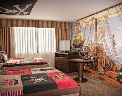 Pirate-themed hotel room with two beds featuring skull and crossbones bedding. A wall mural shows a ship deck, complete with a parrot and sea view.