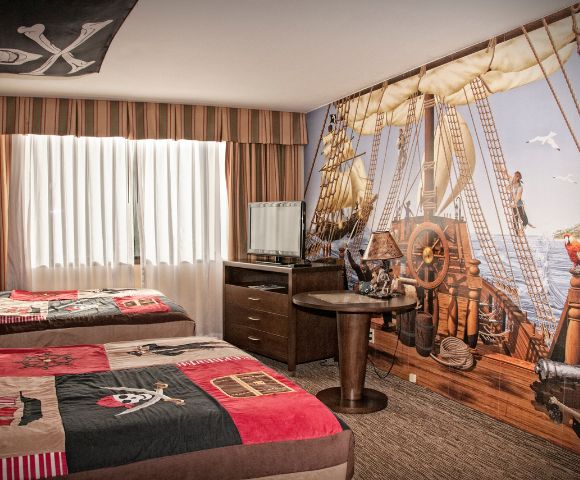 Pirate-themed hotel room with two beds featuring skull and crossbones bedding. A wall mural shows a ship deck, complete with a parrot and sea view.
