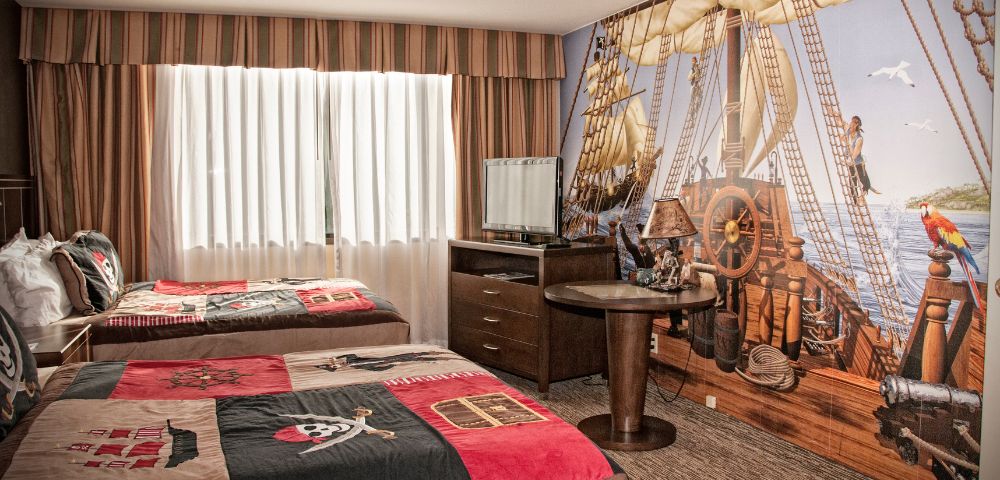 Pirate-themed hotel room with two beds featuring skull and crossbones bedding. A wall mural shows a ship deck, complete with a parrot and sea view.