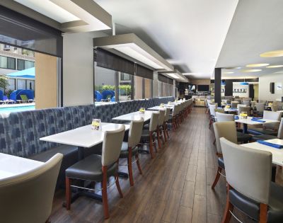 A modern restaurant interior with wooden flooring and large windows overlooking a pool area. Tables are neatly arranged with gray chairs, conveying a welcoming atmosphere.