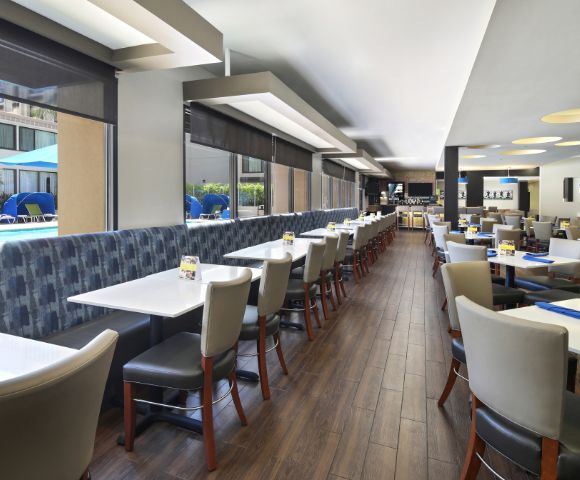 A modern restaurant interior with wooden flooring and large windows overlooking a pool area. Tables are neatly arranged with gray chairs, conveying a welcoming atmosphere.