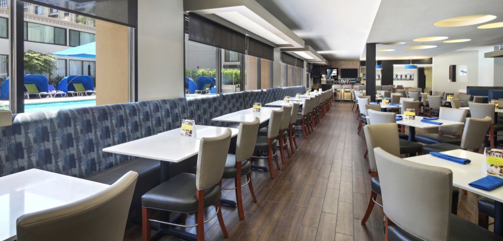 A modern restaurant interior with wooden flooring and large windows overlooking a pool area. Tables are neatly arranged with gray chairs, conveying a welcoming atmosphere.