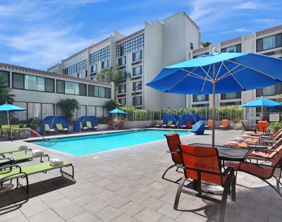Hotel pool area with blue umbrellas and lounge chairs around a clear, rectangular swimming pool. Modern building in the background under a clear sky.