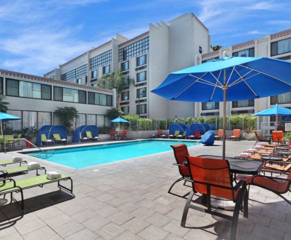 Hotel pool area with blue umbrellas and lounge chairs around a clear, rectangular swimming pool. Modern building in the background under a clear sky.