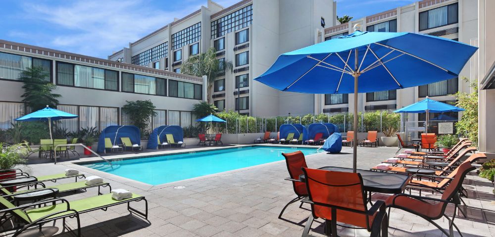 Hotel pool area with blue umbrellas and lounge chairs around a clear, rectangular swimming pool. Modern building in the background under a clear sky.