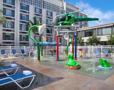 Children's splash pad with helicopter and flower water features, bright colors, surrounded by lounge chairs and palm trees, creating a fun, relaxed vibe.