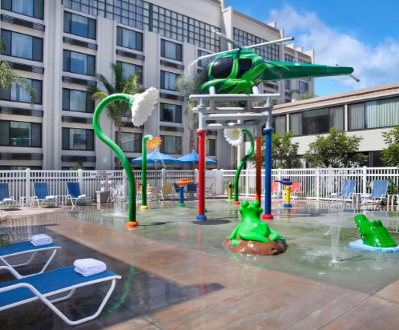 Children's splash pad with helicopter and flower water features, bright colors, surrounded by lounge chairs and palm trees, creating a fun, relaxed vibe.