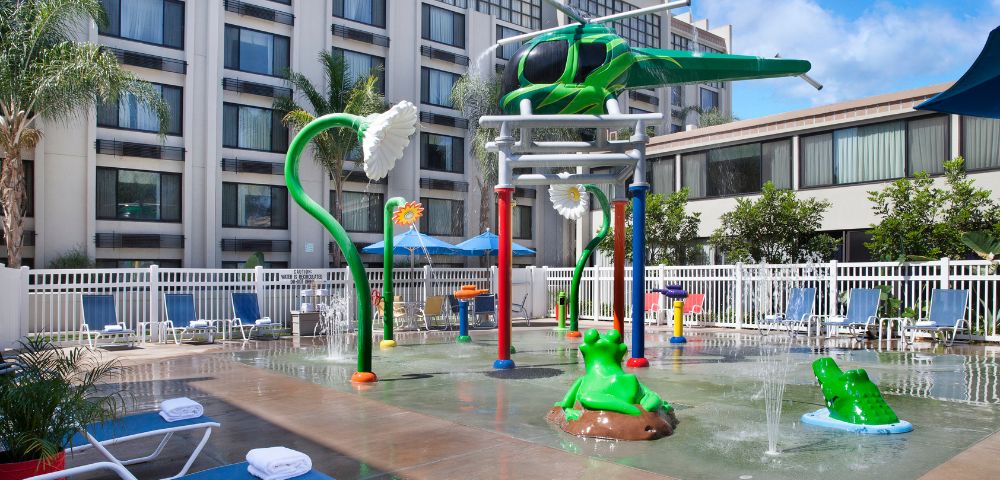 Children's splash pad with helicopter and flower water features, bright colors, surrounded by lounge chairs and palm trees, creating a fun, relaxed vibe.