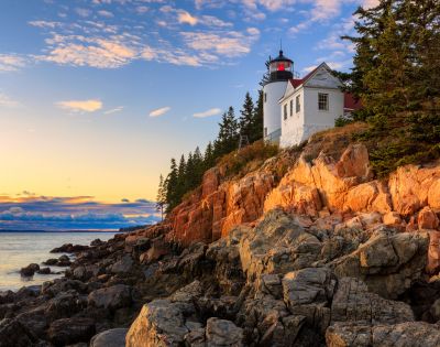 A lighthouse on a rocky cliff is bathed in warm sunset light. Tall pine trees surround it, and the calm sea reflects the colorful sky, creating a serene atmosphere.