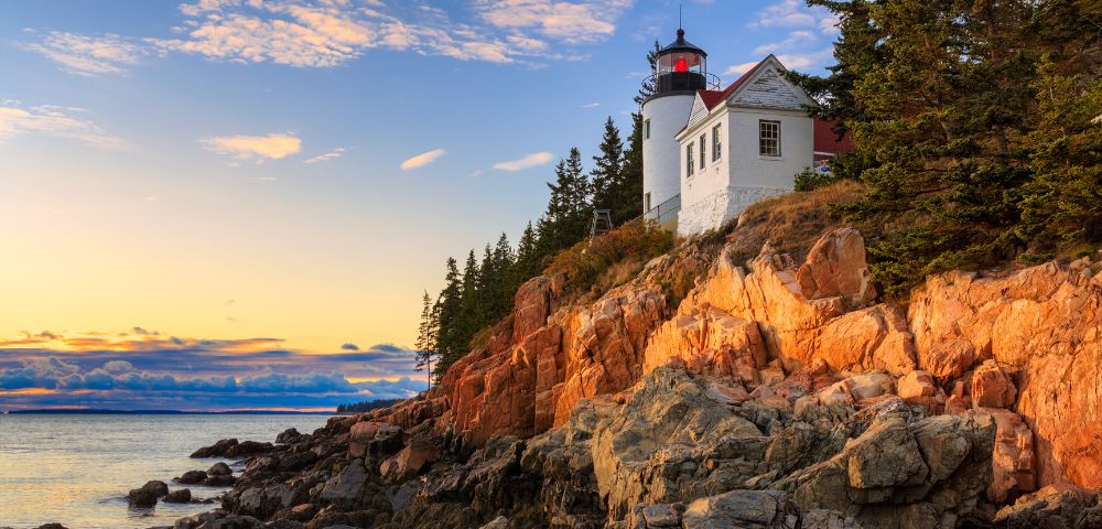 A lighthouse on a rocky cliff is bathed in warm sunset light. Tall pine trees surround it, and the calm sea reflects the colorful sky, creating a serene atmosphere.