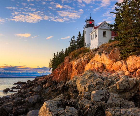 A lighthouse on a rocky cliff is bathed in warm sunset light. Tall pine trees surround it, and the calm sea reflects the colorful sky, creating a serene atmosphere.