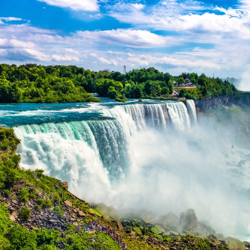 Vibrant view of Niagara Falls, with powerful cascading water, mist rising into a blue sky, and lush green trees lining the background, evoking awe.