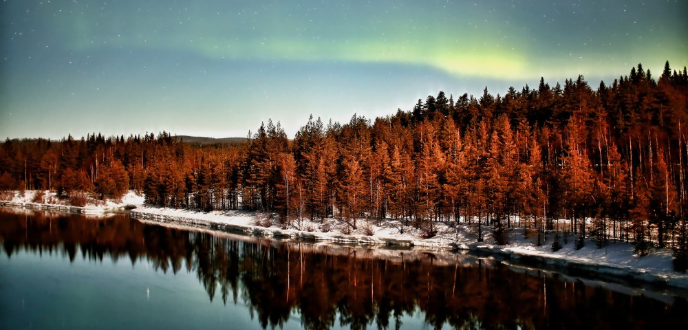 Northern lights glow above a forest reflected in a calm river. Snow lines the riverbank, while stars twinkle in the clear night sky, evoking serenity.