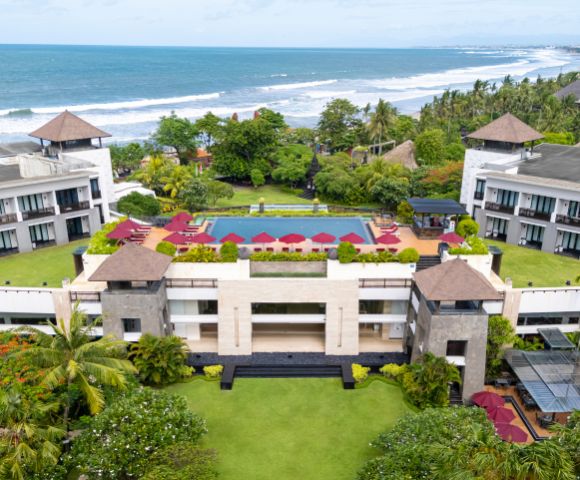 Aerial view of a coastal resort with a central rectangular pool surrounded by red umbrellas. Lush greenery and the ocean enhance its tranquil setting.