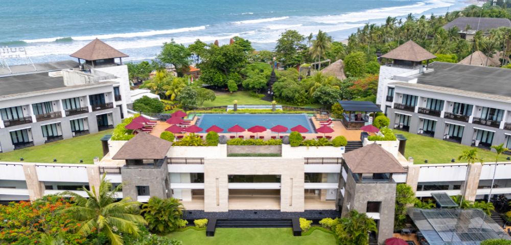 Aerial view of a coastal resort with a central rectangular pool surrounded by red umbrellas. Lush greenery and the ocean enhance its tranquil setting.