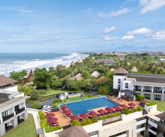 Aerial view of a seaside resort with a large pool surrounded by red umbrellas and lush greenery, overlooking a beach with rolling waves under a blue sky.