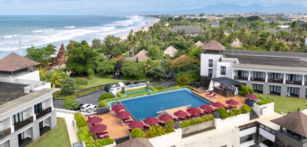 Aerial view of a seaside resort with a large pool surrounded by red umbrellas and lush greenery, overlooking a beach with rolling waves under a blue sky.