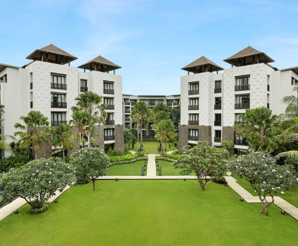 Luxurious resort courtyard with manicured lawns, palm trees, and white buildings under clear blue skies. The scene conveys serenity and elegance.