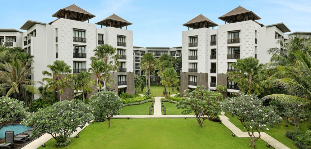 Luxurious resort courtyard with manicured lawns, palm trees, and white buildings under clear blue skies. The scene conveys serenity and elegance.