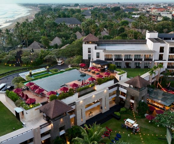 Aerial view of a luxurious beachfront resort with a central pool surrounded by red umbrellas, lush greenery, and a scenic beach in the background.