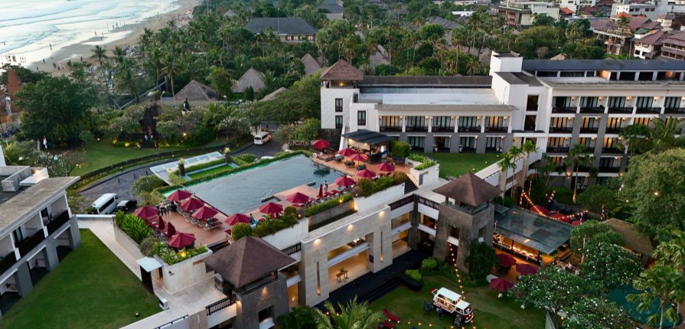 Aerial view of a luxurious beachfront resort with a central pool surrounded by red umbrellas, lush greenery, and a scenic beach in the background.