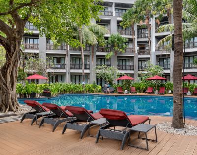 Sunlit hotel pool area with red loungers and umbrellas, surrounded by lush greenery and tall palm trees. A serene, inviting atmosphere.