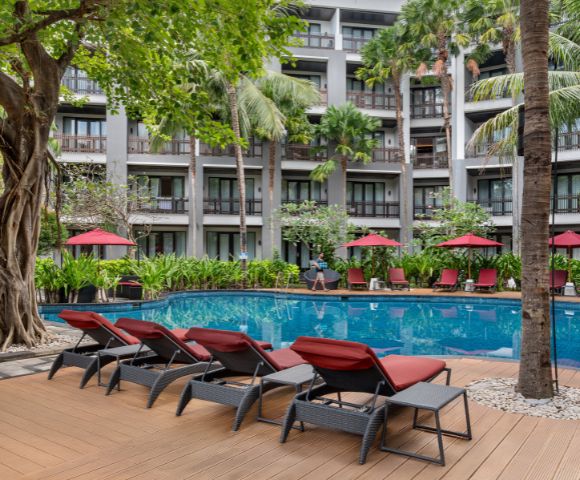 Sunlit hotel pool area with red loungers and umbrellas, surrounded by lush greenery and tall palm trees. A serene, inviting atmosphere.