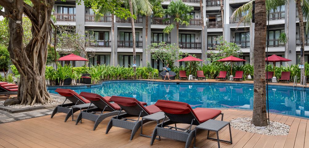 Sunlit hotel pool area with red loungers and umbrellas, surrounded by lush greenery and tall palm trees. A serene, inviting atmosphere.