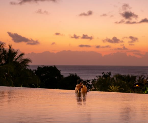 A couple embraces in an infinity pool at sunset, surrounded by tropical trees. The sky is a gradient of orange and pink, creating a romantic ambiance.
