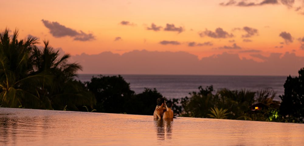 A couple embraces in an infinity pool at sunset, surrounded by tropical trees. The sky is a gradient of orange and pink, creating a romantic ambiance.