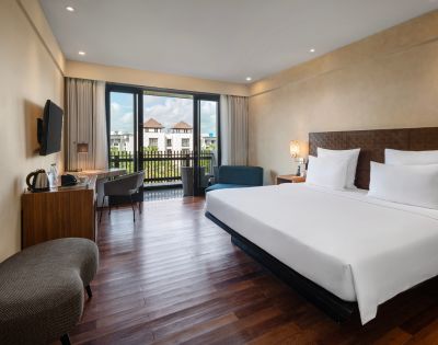 Modern hotel room with large bed, white linens, wooden floor, and a window view of buildings with red roofs. Includes a desk, chair, and cozy seating.