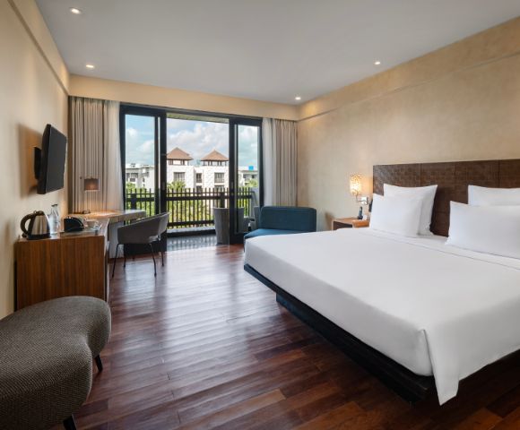 Modern hotel room with large bed, white linens, wooden floor, and a window view of buildings with red roofs. Includes a desk, chair, and cozy seating.