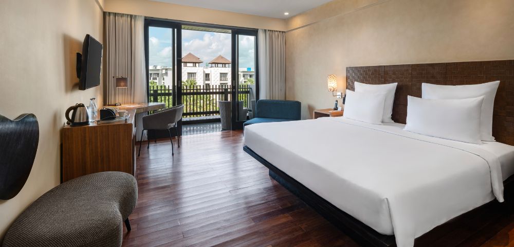 Modern hotel room with large bed, white linens, wooden floor, and a window view of buildings with red roofs. Includes a desk, chair, and cozy seating.