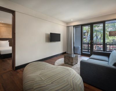 Modern hotel suite with wooden floors, featuring a cozy living area with a gray sofa, TV, and pouf. Bedroom with a plush bed is visible; balcony with palm tree view.